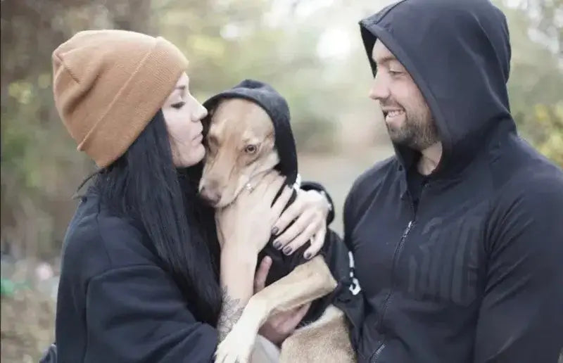 Couple in hoodies cuddling a dog wearing an Adidog Hooded Sweatshirt in a cozy outdoor setting.