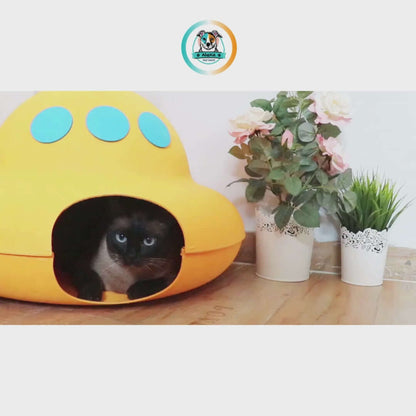 Cat peeking out from an orange INKZOO cat cave with blue spots, surrounded by plants.